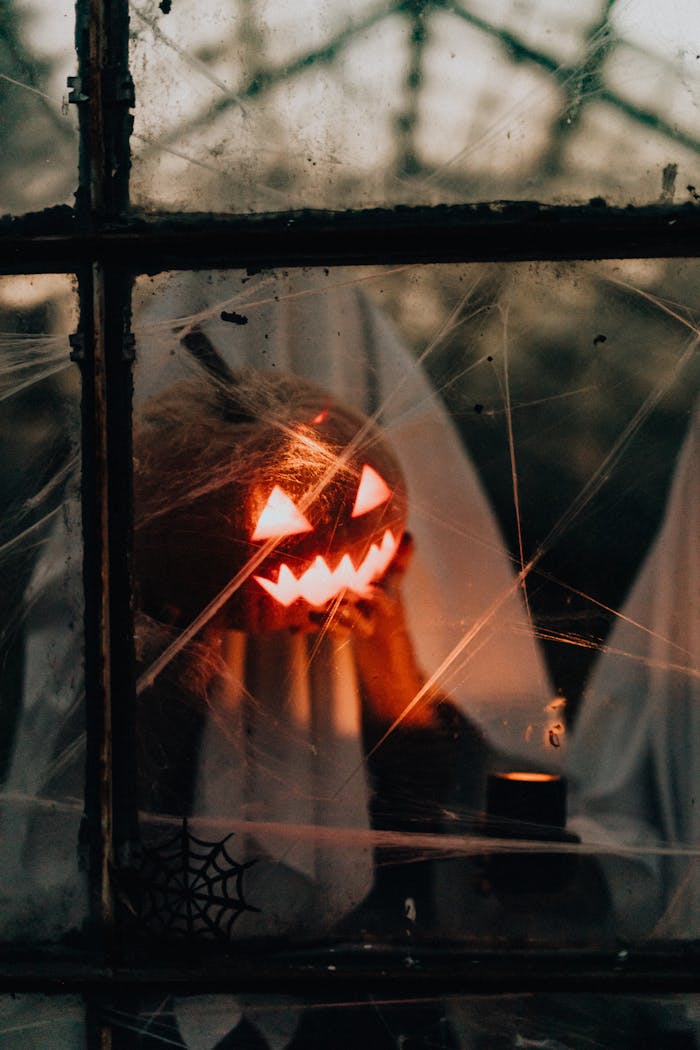 A haunting Halloween scene with illuminated jack-o-lanterns viewed through a spiderwebbed window.