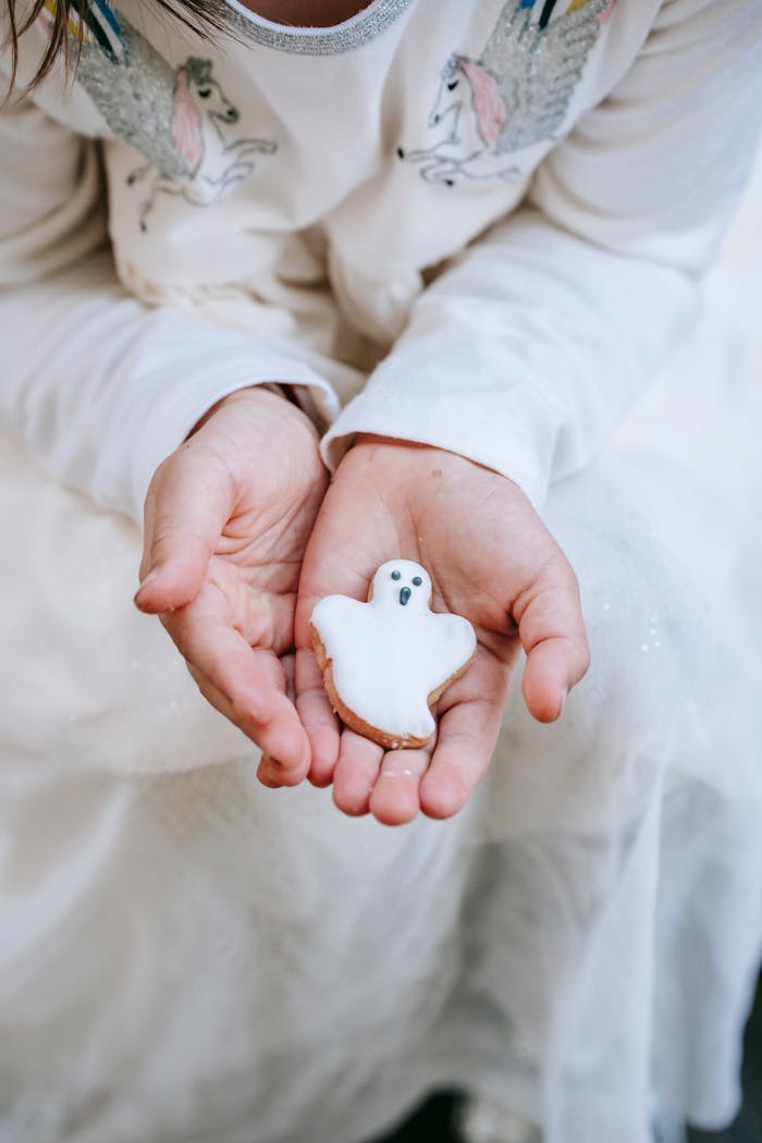 From above crop girl wearing stylish white dress showing sweet gingerbread ghost cookie on hands during Halloween celebration