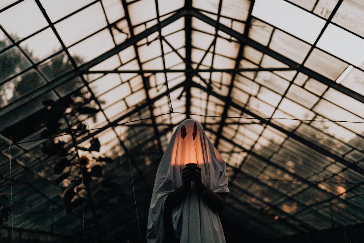 Mysterious figure in ghost costume holding lantern inside a dimly lit greenhouse.