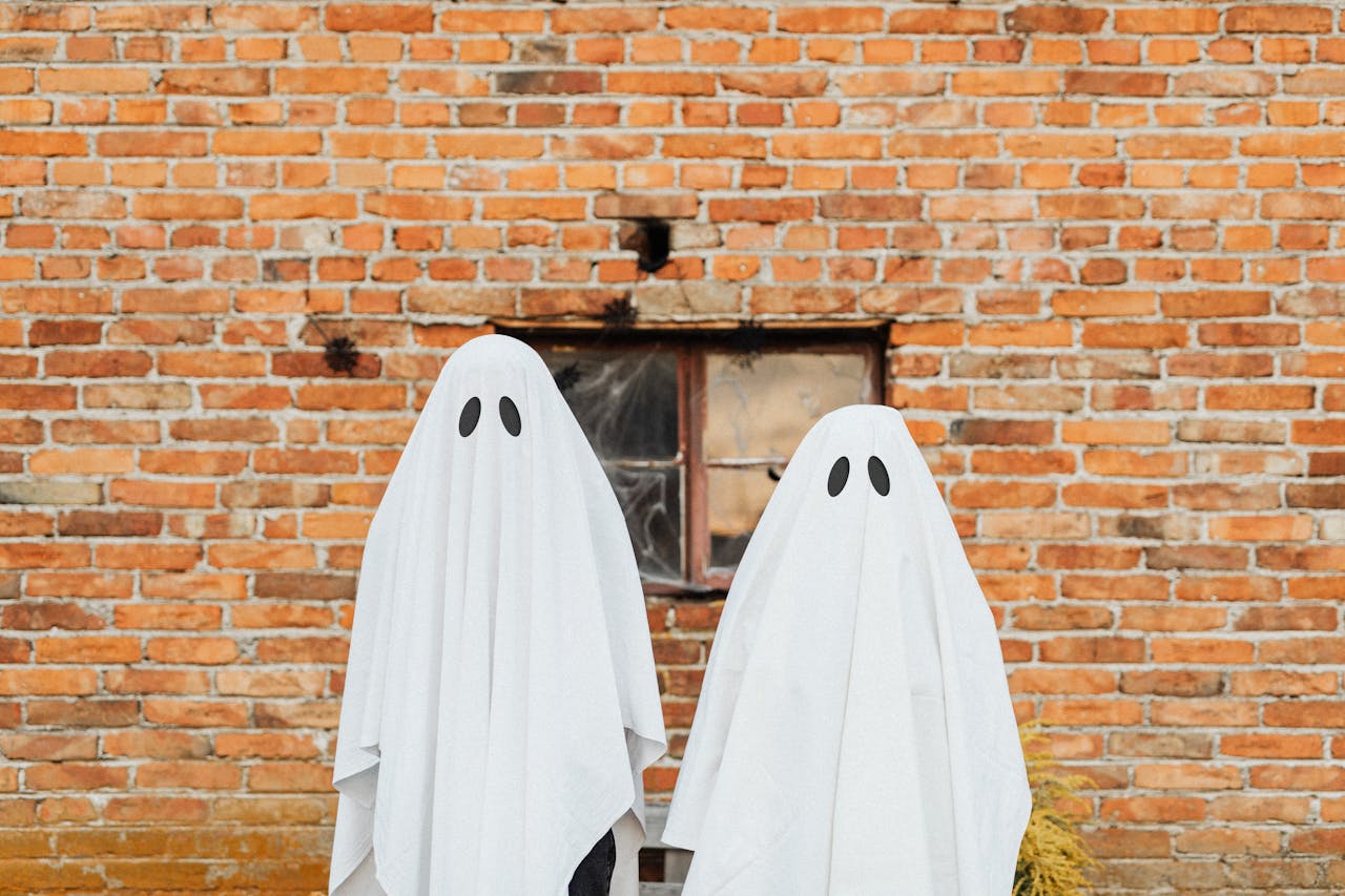 Two ghostly figures in white sheets stand against a rustic brick wall for Halloween.
