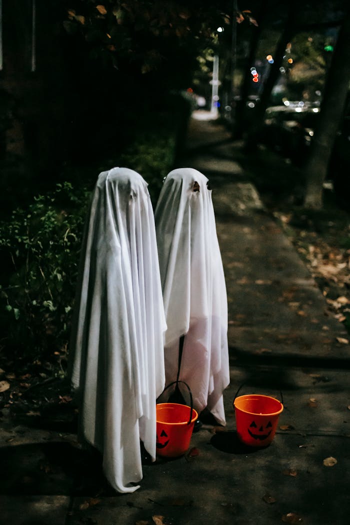 Full body side view of anonymous kids wearing white ghost costumes standing on sidewalk with orange buckets while playing trick or treat in evening