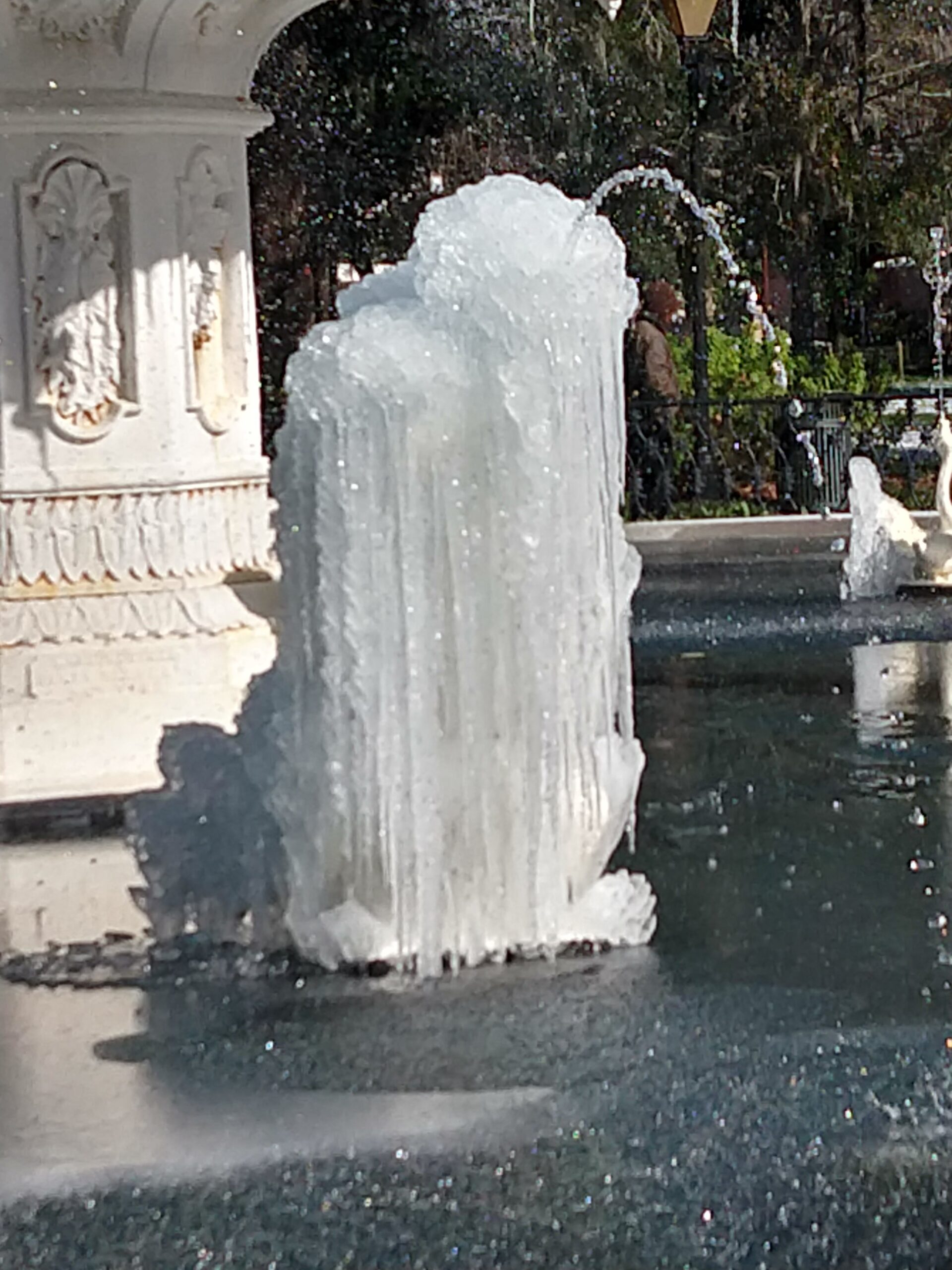 A Forsyth Park fountain statue completely encased in a thick column of ice during January 2018 — water still actively shooting from the top of the ice column, proving the pumps never stopped